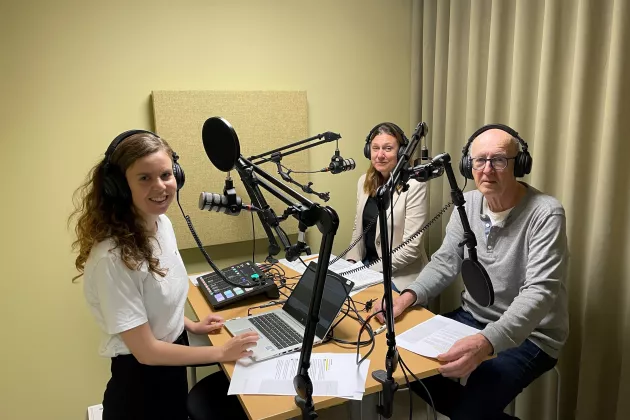 Three persons are standing in a studio, recording a podcast
